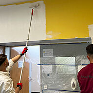 Ein Schüler im Maler-Overall trägt weiße Farbe auf einer gelben Wand auf.