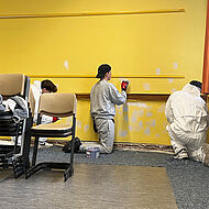 Drei Schüler in einem Klassenzimmer beim Abschleifen von frisch verputzten Stellen.